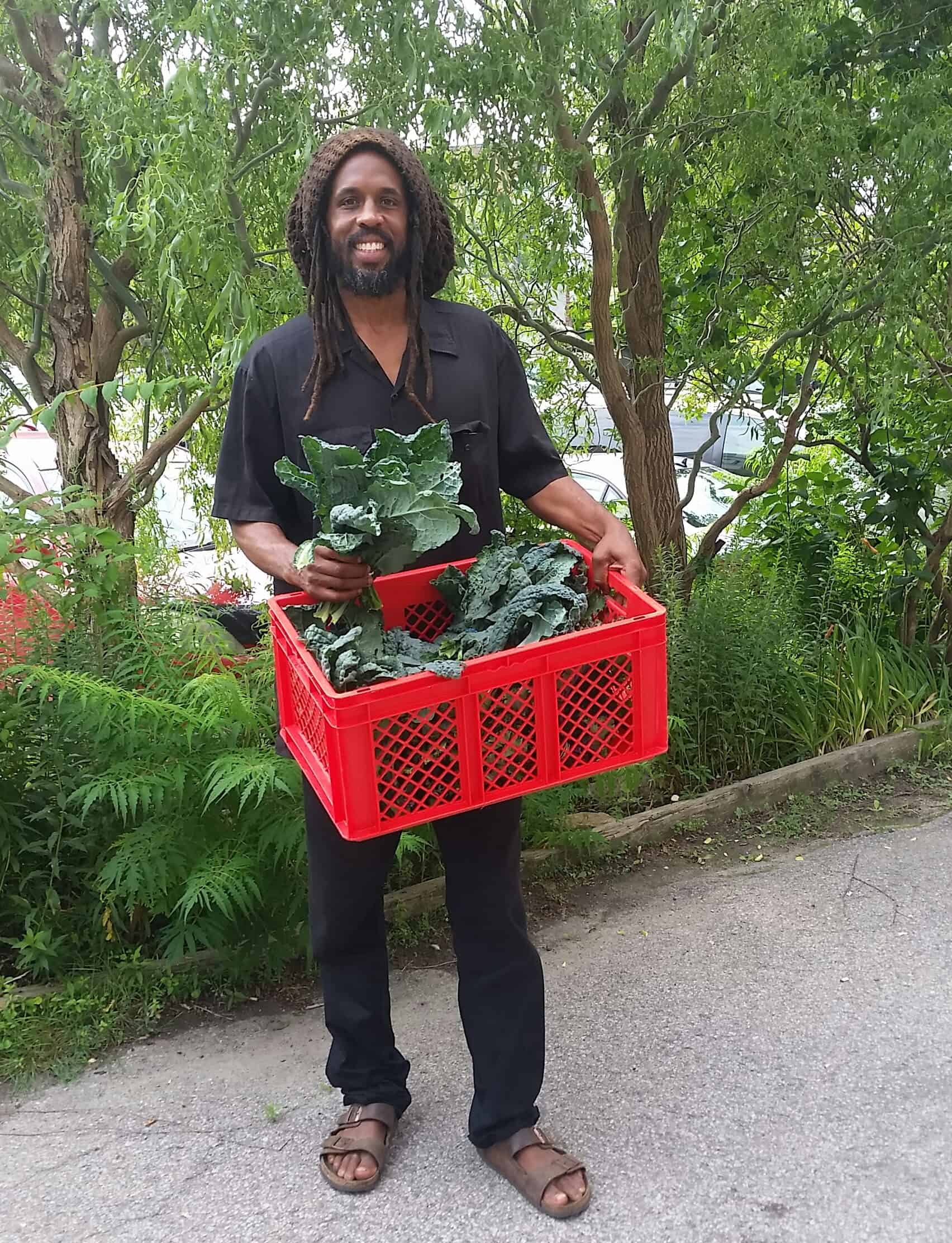 Shabaka standing tall and smiling with beard, dreadlocks, and a crate of crisp kale