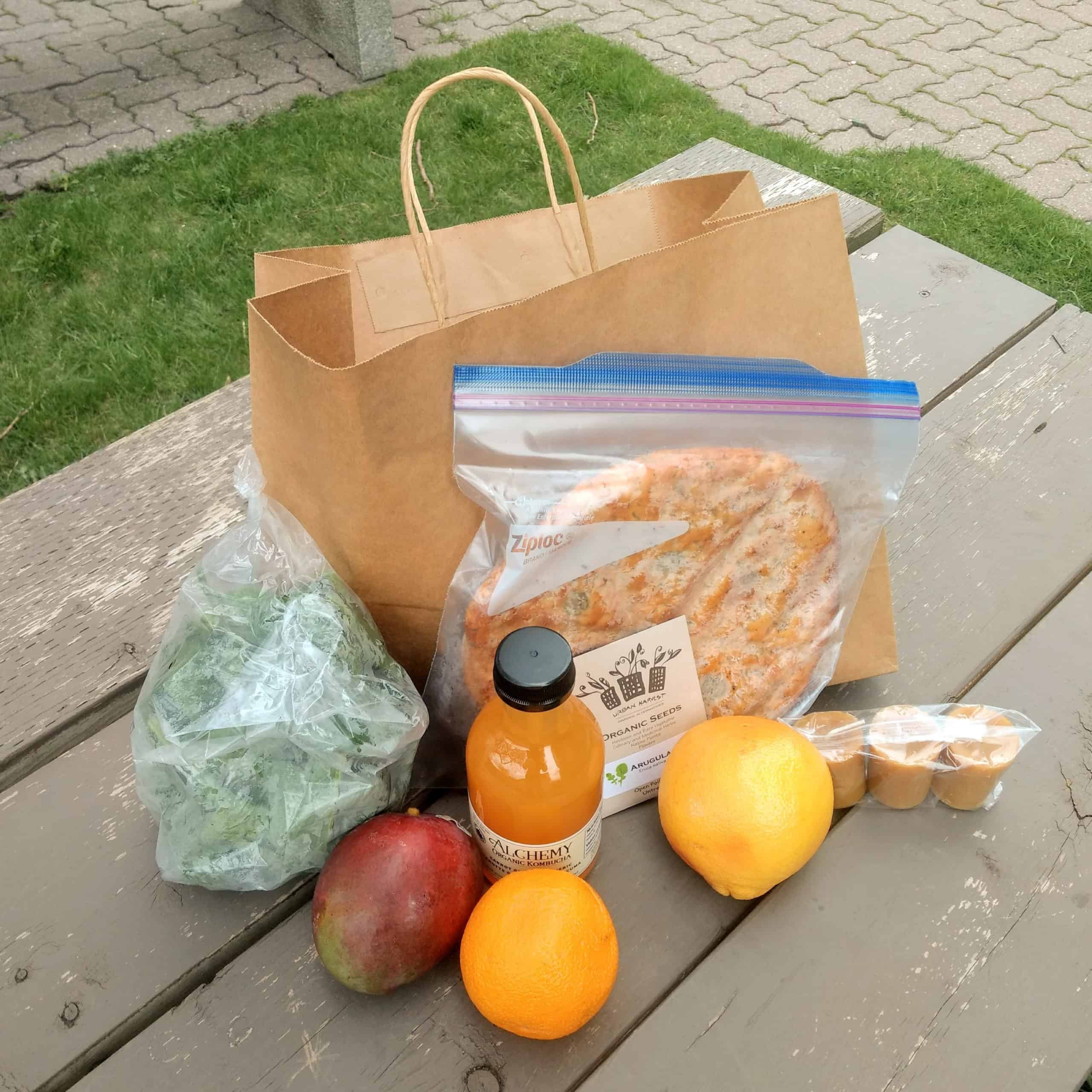 A bag on a picnic table surrounded by fruit, green, kombucha, seeds, bread, and beeswax candles